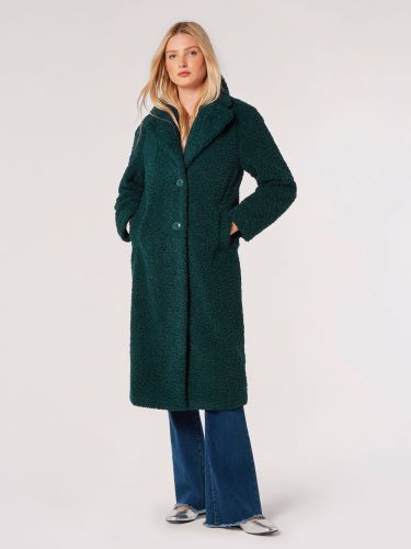 A person stands against a plain background, wearing an Apricot Boucle textured green coat with a collar and buttons down the front, paired with blue jeans and silver shoes